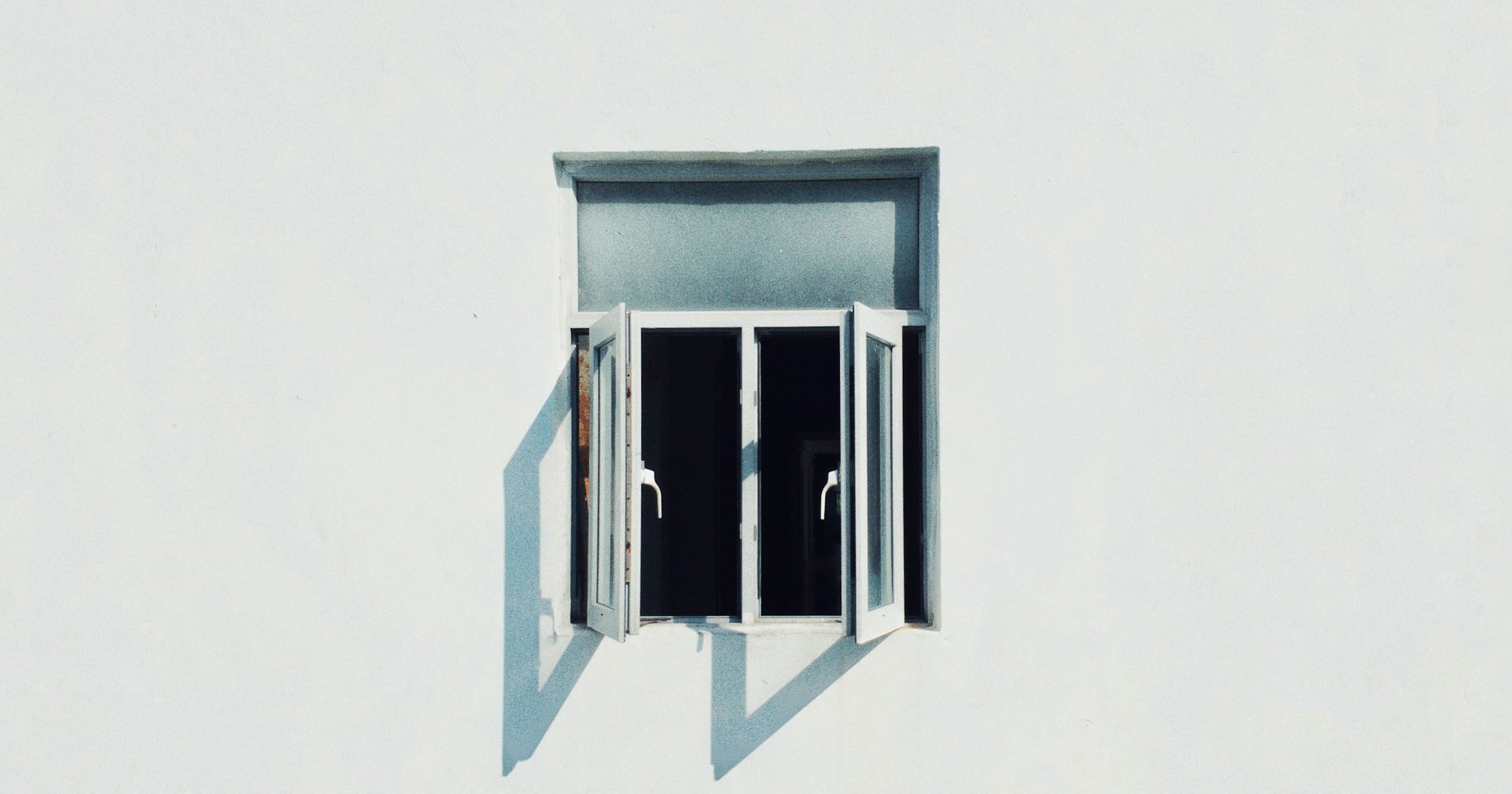 An open window in a white wall, with the room behind it in darkness. Photo by Jason Yuen on Unsplash.